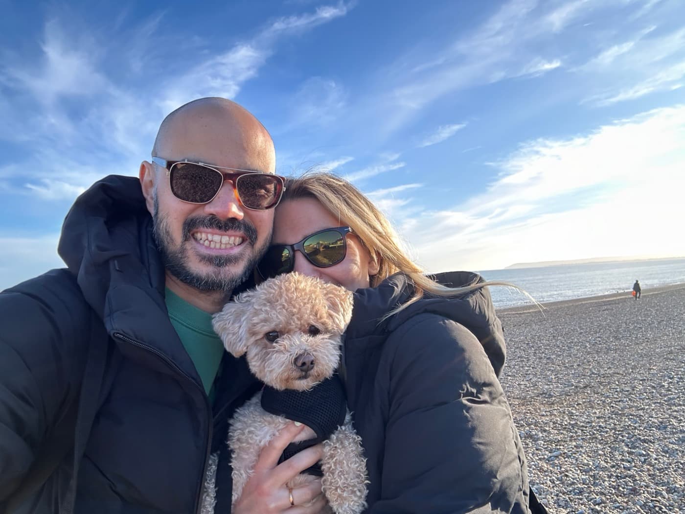 Josh and Debbie smiling on a pebble beach, holding Bobby, a small cream-coloured dog in a black harness